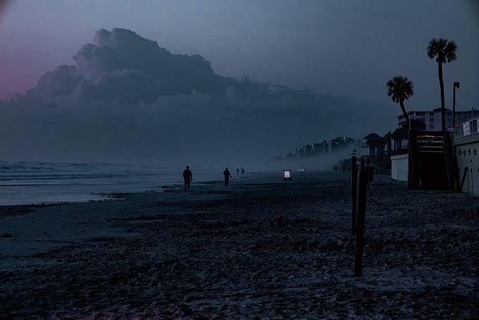 A foggy beach at dusk with two distant figures and a huge cloud bank over the water.