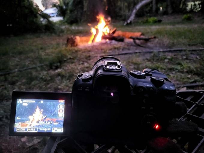 A camera on a tripod filming a campfire at dusk, flame visible on its flip-out screen.