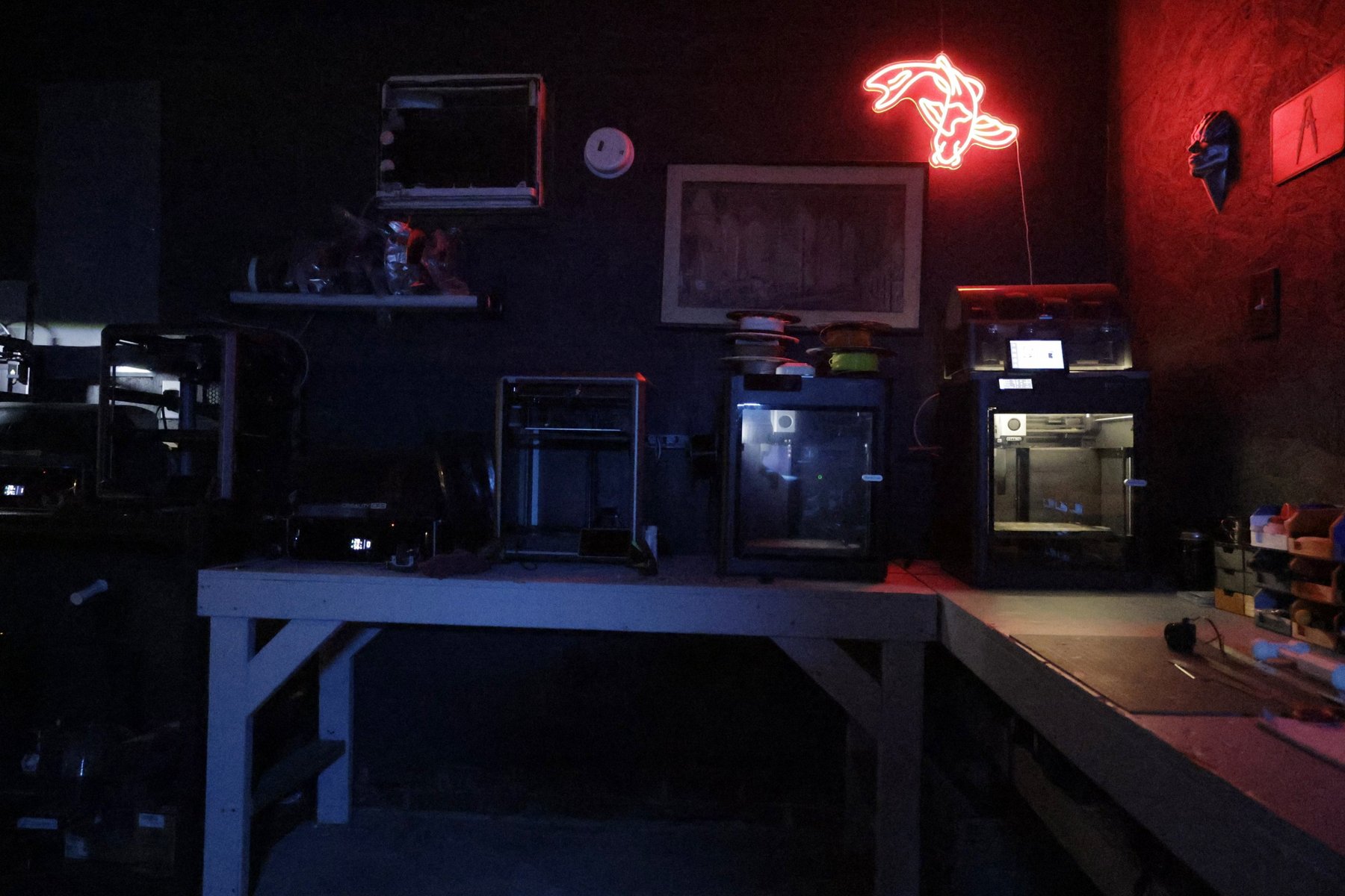 Several enclosed 3D printers in a row on the TacticalTech3D bench, filament spools stacked above them, under a red neon sign.